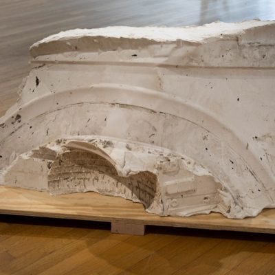 A white, rectangular shaped abstract cast plaster sculpture with an arch impressed in the side and tire treads